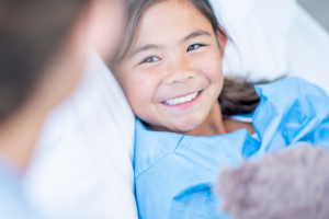a young girl smiling holding a teddy bear on a hospital bed