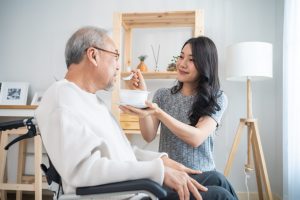 Asian beautiful young daughter support to elderly father on wheelchair. Attractive woman serving and feeding food to older patient grandfather in living room at home. Family Healthcare medical concept